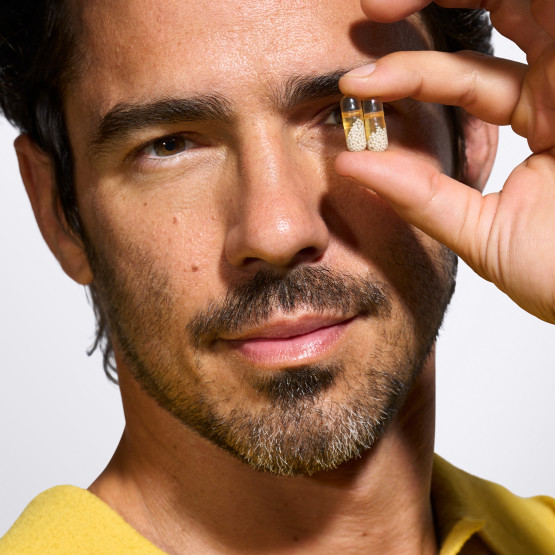 Male model holding two capsules in hand in front of left eye.
